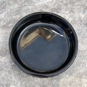 May include: A circular, black ceramic dish with a shiny surface. The dish has a raised edge and a smooth inside. The surface reflects light, and the dish appears empty. The dish is on a grey surface.