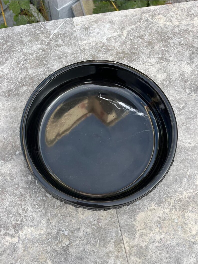 May include: A circular, black ceramic dish with a shiny surface. The dish has a raised edge and a smooth inside. The surface reflects light, and the dish appears empty. The dish is on a grey surface.