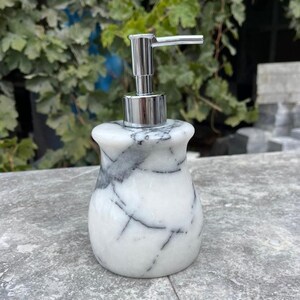 Puede incluir: Un dispensador de jab&oacute;n de m&aacute;rmol blanco con vetas grises. El dispensador tiene una bomba cromada y una base redondeada. El fondo presenta follaje verde y una superficie gris.