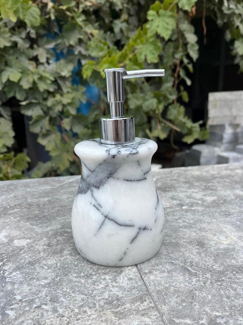 Puede incluir: Un dispensador de jab&oacute;n de m&aacute;rmol blanco con vetas grises. El dispensador tiene una bomba cromada y una base redondeada. El fondo presenta follaje verde y una superficie gris.