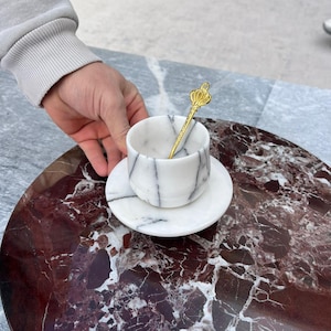 Op de afbeelding: Een marmeren kop en schotel set met zwarte adering, vergezeld van een goudkleurige lepel met een kroonontwerp. De set staat op een rond, roodbruin marmeren oppervlak, wat een luxe uitstraling geeft.