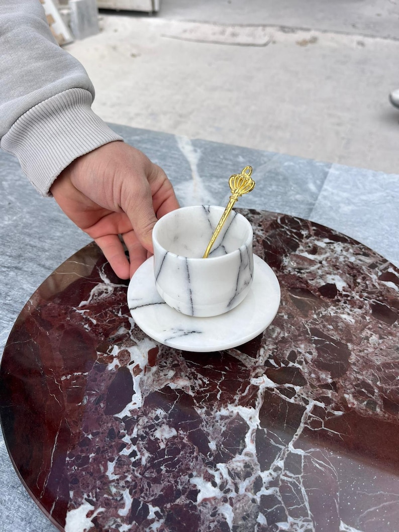 Op de afbeelding: Een marmeren kop en schotel set met zwarte adering, vergezeld van een goudkleurige lepel met een kroonontwerp. De set staat op een rond, roodbruin marmeren oppervlak, wat een luxe uitstraling geeft.