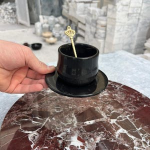 Op de afbeelding: Een zwarte keramische espressokop en schotelset met een goudkleurige kroonvormige lepel. De kop en schotel staan op een rood en wit marmeren oppervlak. De achtergrond toont een werkplaats met marmeren blokken.