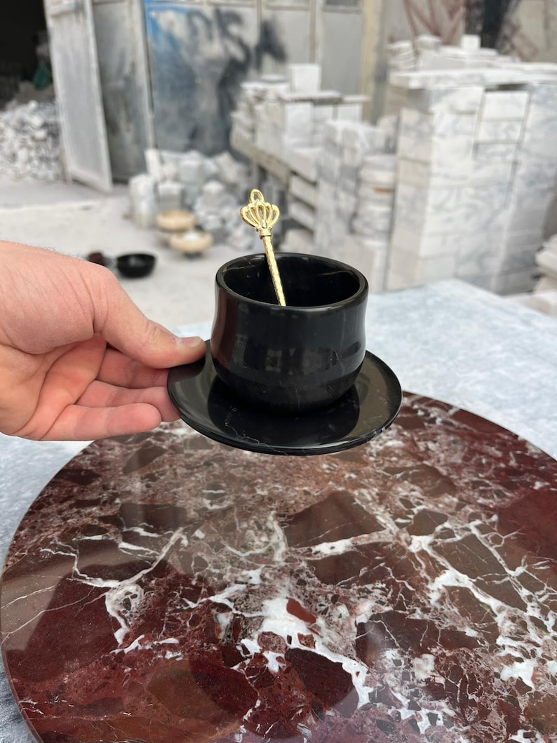 Op de afbeelding: Een zwarte keramische espressokop en schotelset met een goudkleurige kroonvormige lepel. De kop en schotel staan op een rood en wit marmeren oppervlak. De achtergrond toont een werkplaats met marmeren blokken.