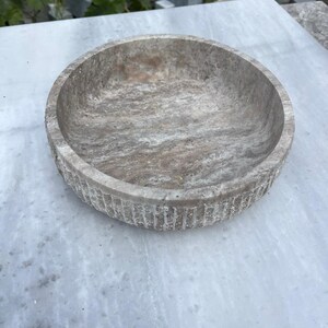 May include: A round, stone bowl with a textured, marbled appearance. The bowl is a light gray color with darker gray and brown veining. The rim has a carved, vertical grooved pattern. The bowl is sitting on a light gray surface.