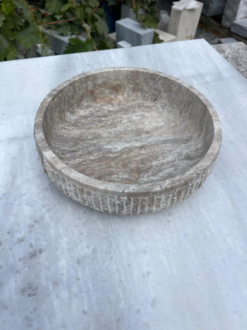 May include: A round, stone bowl with a textured, marbled appearance. The bowl is a light gray color with darker gray and brown veining. The rim has a carved, vertical grooved pattern. The bowl is sitting on a light gray surface.