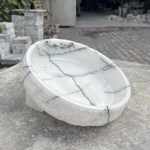 May include: A white marble bowl with black veining. The bowl has a rounded shape and sits on a small, rectangular base. The exterior has a rough, textured finish, while the interior is smooth. The bowl is likely for decorative or functional use.