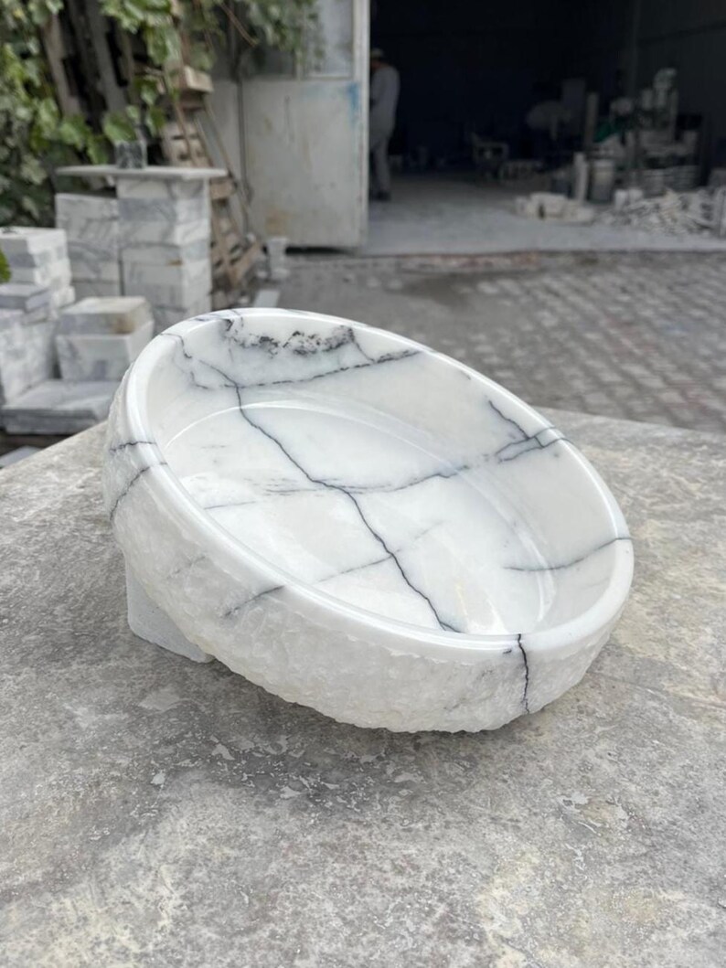 May include: A white marble bowl with black veining. The bowl has a rounded shape and sits on a small, rectangular base. The exterior has a rough, textured finish, while the interior is smooth. The bowl is likely for decorative or functional use.