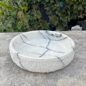 May include: A white marble bowl with a textured exterior and a smooth interior. The bowl has black veining across its surface. The round bowl is displayed on a grey surface.