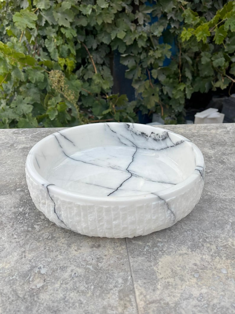 May include: A white marble bowl with a textured exterior and a smooth interior. The bowl has black veining across its surface. The round bowl is displayed on a grey surface.