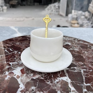 Op de afbeelding: Een witte keramische espressokop en schotelset met een goudkleurige lepel. De lepel heeft een kroonontwerp op het handvat. De set staat op een rood-bruin marmeren oppervlak.