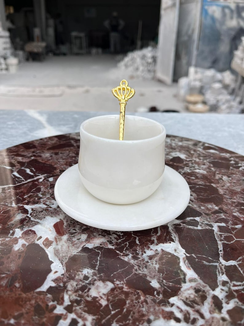 Op de afbeelding: Een witte keramische espressokop en schotelset met een goudkleurige lepel. De lepel heeft een kroonontwerp op het handvat. De set staat op een rood-bruin marmeren oppervlak.