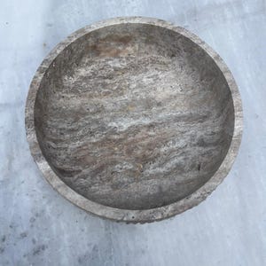 May include: A round, stone bowl with a textured surface. The bowl has a light gray and brown color scheme, with a rough, natural appearance. The bowl's rim has a slightly uneven edge.