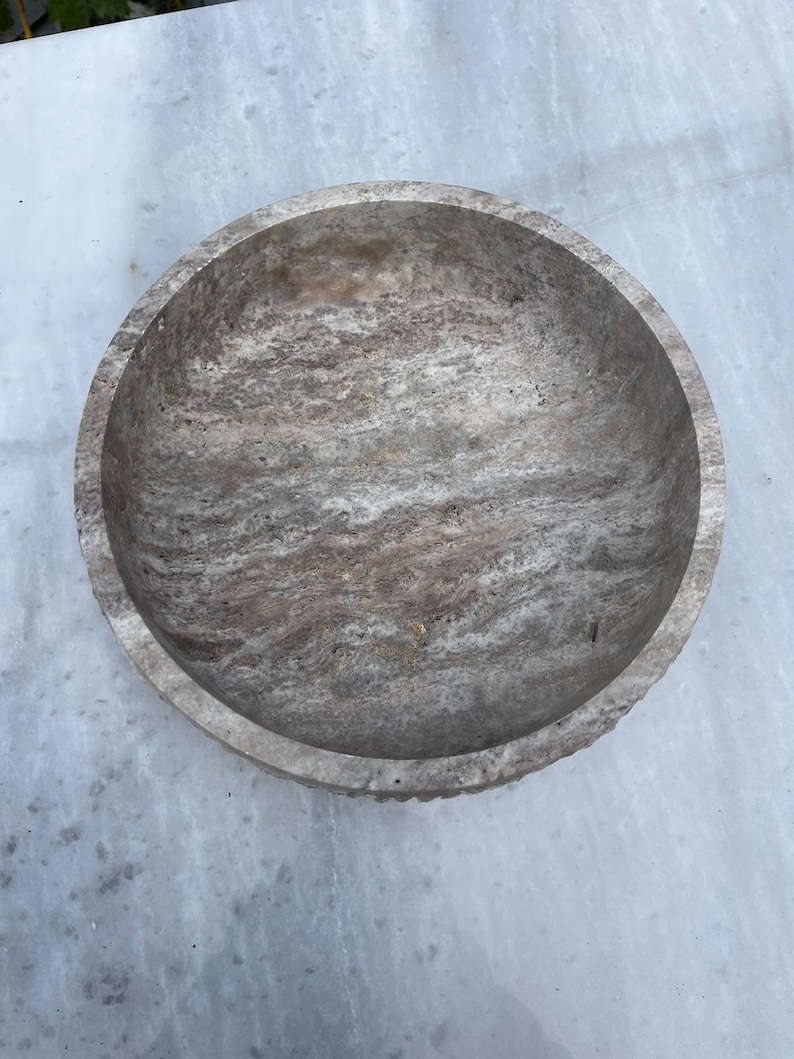 May include: A round, stone bowl with a textured surface. The bowl has a light gray and brown color scheme, with a rough, natural appearance. The bowl's rim has a slightly uneven edge.