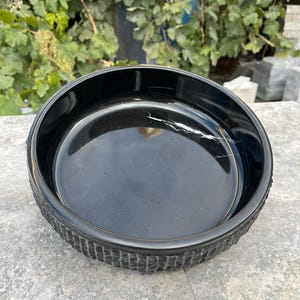 May include: A black pet bowl with a textured, ridged exterior. The interior is smooth and glossy. The bowl is round and sits on a grey surface, with green foliage in the background.