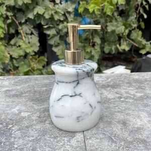 Puede incluir: Un dispensador de jab&oacute;n de m&aacute;rmol blanco con vetas negras y una bomba dorada. El dispensador tiene forma redondeada y se apoya sobre una superficie gris, con follaje verde al fondo. Accesorio de ba&ntilde;o.
