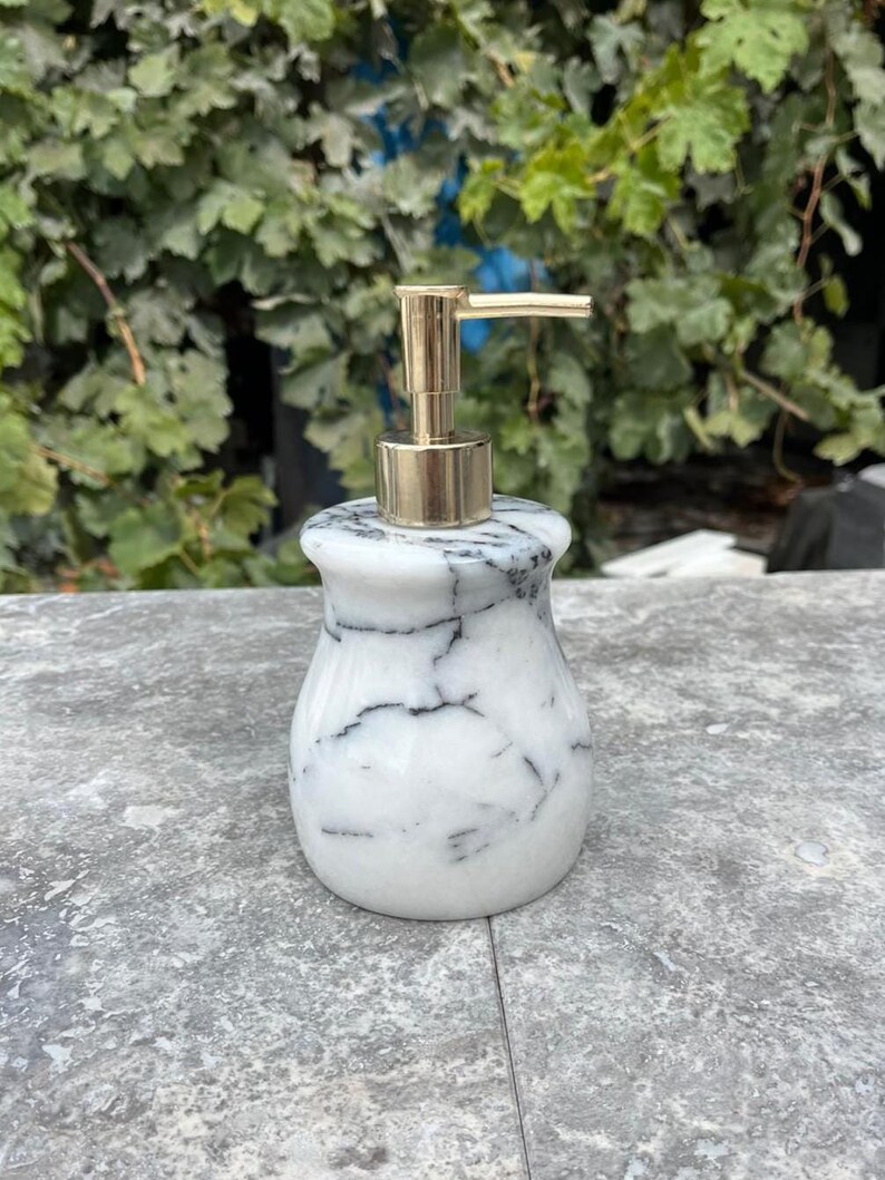 Puede incluir: Un dispensador de jab&oacute;n de m&aacute;rmol blanco con vetas negras y una bomba dorada. El dispensador tiene forma redondeada y se apoya sobre una superficie gris, con follaje verde al fondo. Accesorio de ba&ntilde;o.