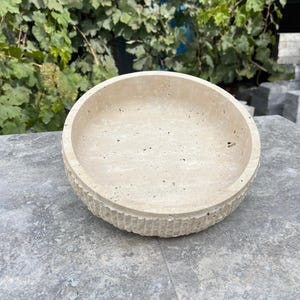 K&ouml;nnte beinhalten: Eine beige, runde Steinschale mit strukturierter Au&szlig;enseite und glatter Innenseite. Die Schale besteht aus einem hellfarbigen Stein und hat ein schlichtes, aber elegantes Design. Die Schale eignet sich f&uuml;r dekorative oder funktionale Zwecke.