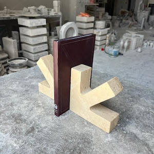 May include: A pair of beige, porous travertine stone bookends with a V-shaped design, supporting a dark maroon hardcover book. The bookends feature a natural, textured surface with visible small holes and a solid, angular construction.