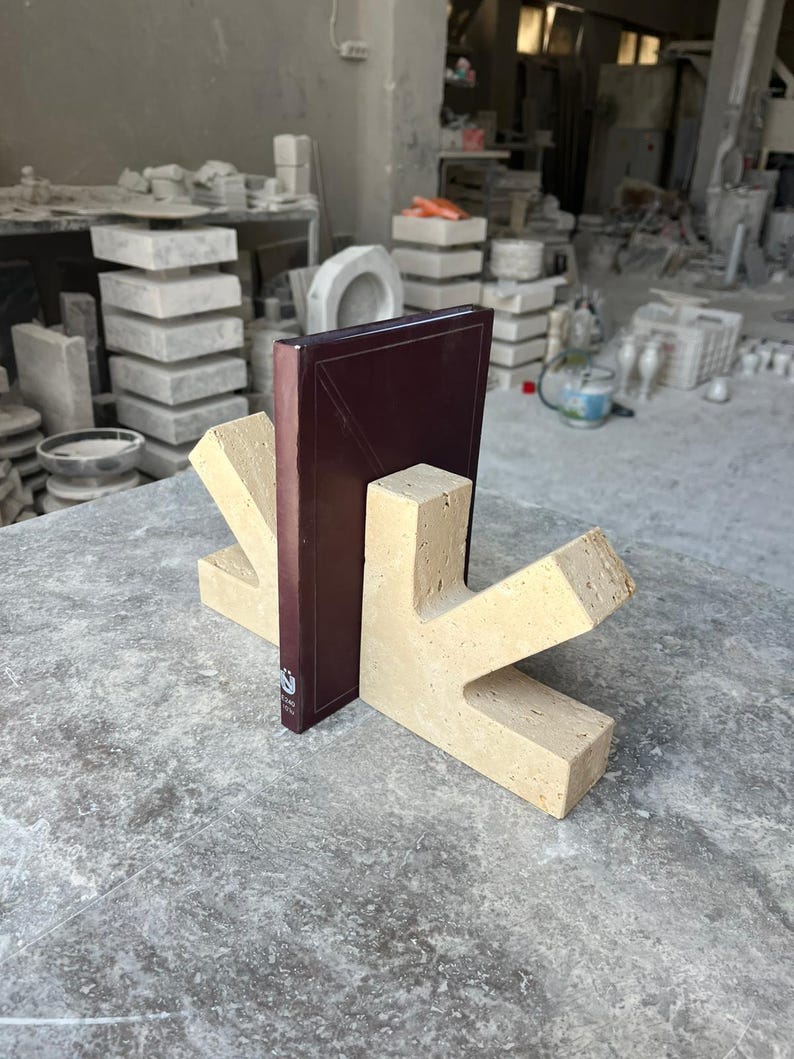 May include: A pair of beige, porous travertine stone bookends with a V-shaped design, supporting a dark maroon hardcover book. The bookends feature a natural, textured surface with visible small holes and a solid, angular construction.