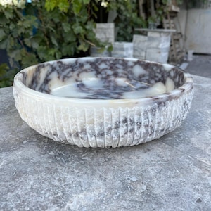 Puede incluir: Un cuenco redondo de piedra tallada con exterior texturizado. El cuenco presenta un patr&oacute;n veteado blanco y marr&oacute;n en el interior. El exterior tiene ranuras verticales. El cuenco est&aacute; sobre una superficie gris.
