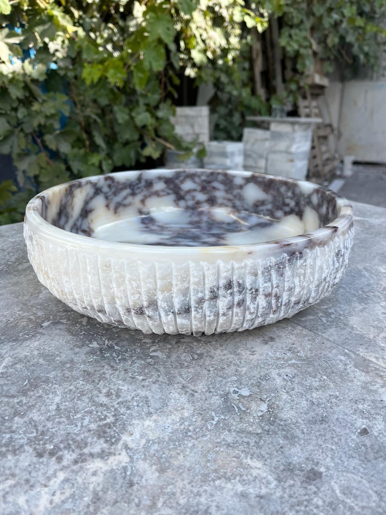 Puede incluir: Un cuenco redondo de piedra tallada con exterior texturizado. El cuenco presenta un patr&oacute;n veteado blanco y marr&oacute;n en el interior. El exterior tiene ranuras verticales. El cuenco est&aacute; sobre una superficie gris.