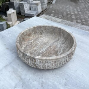 May include: A round, stone bowl with a textured exterior and a smooth interior. The bowl is a neutral color with streaks of gray and brown. The bowl is sitting on a marble surface.