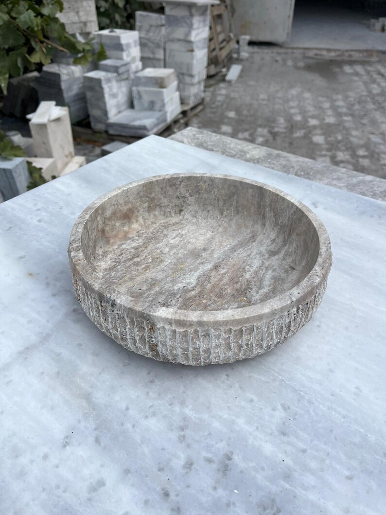 May include: A round, stone bowl with a textured exterior and a smooth interior. The bowl is a neutral color with streaks of gray and brown. The bowl is sitting on a marble surface.