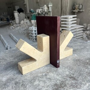 May include: A beige travertine stone bookend with a geometric, angular shape featuring a central vertical slot. A dark maroon book is inserted into the slot, displaying the white text "&Uuml;" and "E240 10M" on its spine. The stone has a porous texture.