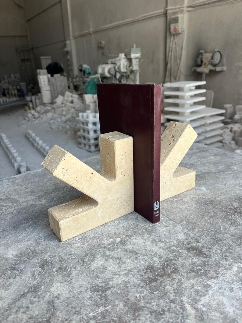 May include: A beige travertine stone bookend with a geometric, angular shape featuring a central vertical slot. A dark maroon book is inserted into the slot, displaying the white text "&Uuml;" and "E240 10M" on its spine. The stone has a porous texture.