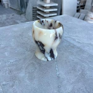 May include: A small, rounded cup carved from polished stone, featuring a creamy off-white base colour with prominent, swirling dark brown and black veining throughout the surface. The vessel has a smooth, glossy finish and a wide, open top.