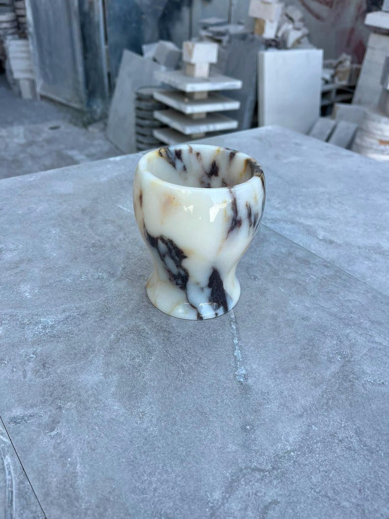 May include: A small, rounded cup carved from polished stone, featuring a creamy off-white base colour with prominent, swirling dark brown and black veining throughout the surface. The vessel has a smooth, glossy finish and a wide, open top.