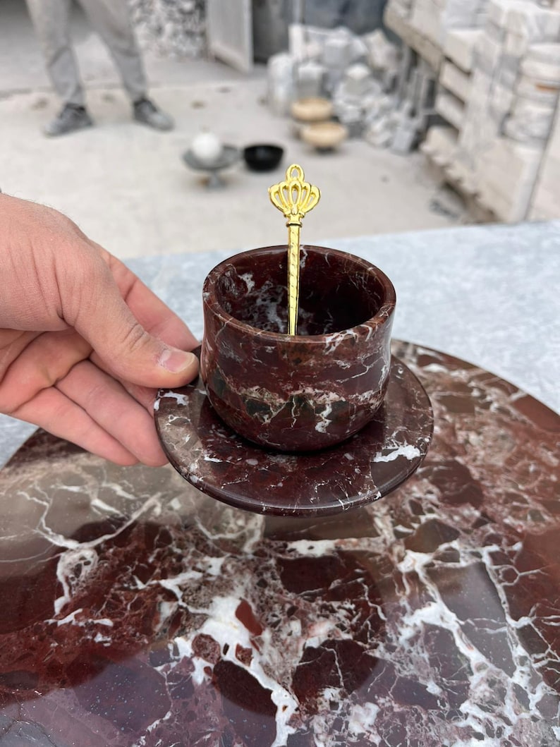 Op de afbeelding: Een rode marmeren kop en schotel set met witte adering. Een goudkleurige lepel met een kroonontwerp rust in de beker. De set wordt gepresenteerd op een bijpassend marmeren oppervlak, dat de patronen van de natuursteen laat zien.