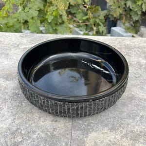 May include: A black, circular bowl with a textured, vertically lined pattern around the sides. The bowl has a glossy finish and sits on a grey surface. The bowl's interior reflects light, showing a subtle white streak.