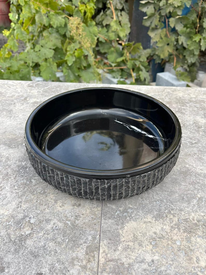 May include: A black, circular bowl with a textured, vertically lined pattern around the sides. The bowl has a glossy finish and sits on a grey surface. The bowl's interior reflects light, showing a subtle white streak.