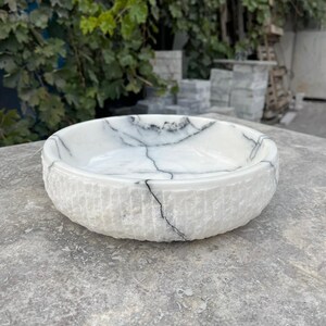 May include: A white marble bowl with black veining. The bowl has a textured, ridged exterior and a smooth interior. The bowl is round and sits on a grey surface. The background features green foliage and a stone structure.