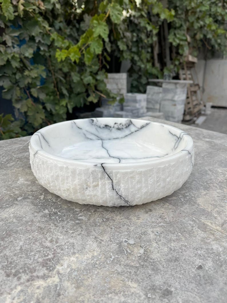 May include: A white marble bowl with black veining. The bowl has a textured, ridged exterior and a smooth interior. The bowl is round and sits on a grey surface. The background features green foliage and a stone structure.