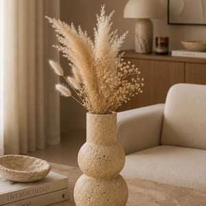 Puede incluir: Un jarr&oacute;n de piedra travertino de color beige, con textura porosa y forma de doble bulbo, que contiene un arreglo de flores secas compuesto por plumas de hierba de la pampa, colas de conejo esponjosas y peque&ntilde;as flores blancas.