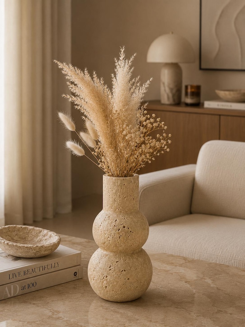 Puede incluir: Un jarr&oacute;n de piedra travertino de color beige, con textura porosa y forma de doble bulbo, que contiene un arreglo de flores secas compuesto por plumas de hierba de la pampa, colas de conejo esponjosas y peque&ntilde;as flores blancas.