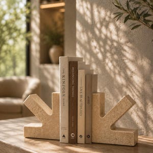 May include: Two beige, porous stone bookends with an angular, geometric shape holding four books titled The Kinfolk Home, Arranging Things, Live Beautifully, and at 100. The bookends feature a textured surface and a symmetrical, stepped design.