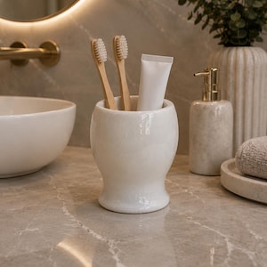 May include: A white ceramic tumbler with a smooth, rounded base and a flared rim. Inside the tumbler are two bamboo toothbrushes with light-coloured bristles and a plain white tube of toothpaste, all standing upright in the centre of the container.
