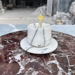 Op de afbeelding: Een witte marmeren espressokop en schotel set met zwarte adering. Een goudkleurige lepel met een kroonontwerp rust in de kop. De set wordt gepresenteerd op een rood en wit marmeren oppervlak, wat een luxe esthetiek laat zien.