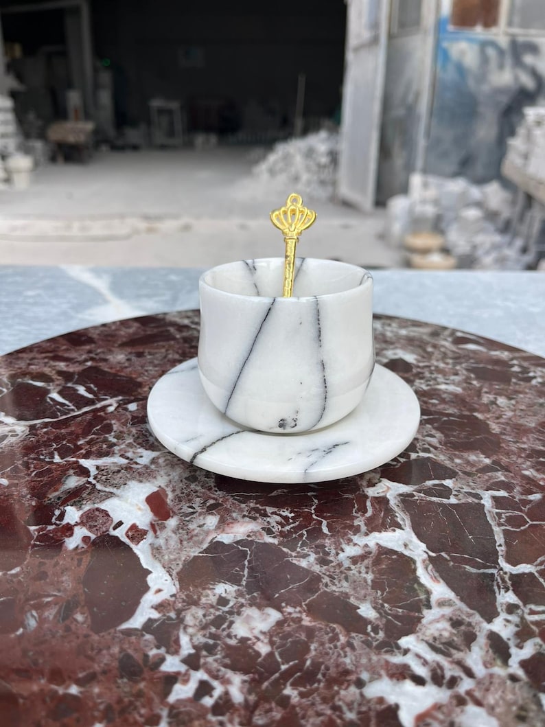 Op de afbeelding: Een witte marmeren espressokop en schotel set met zwarte adering. Een goudkleurige lepel met een kroonontwerp rust in de kop. De set wordt gepresenteerd op een rood en wit marmeren oppervlak, wat een luxe esthetiek laat zien.