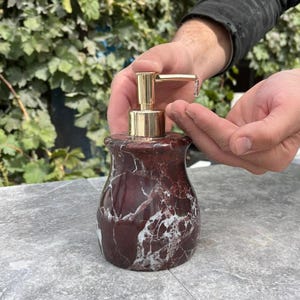Puede incluir: Un dispensador de jab&oacute;n de m&aacute;rmol con una bomba dorada. El dispensador es de color rojo oscuro con vetas blancas. El dispensador est&aacute; sobre una superficie gris.