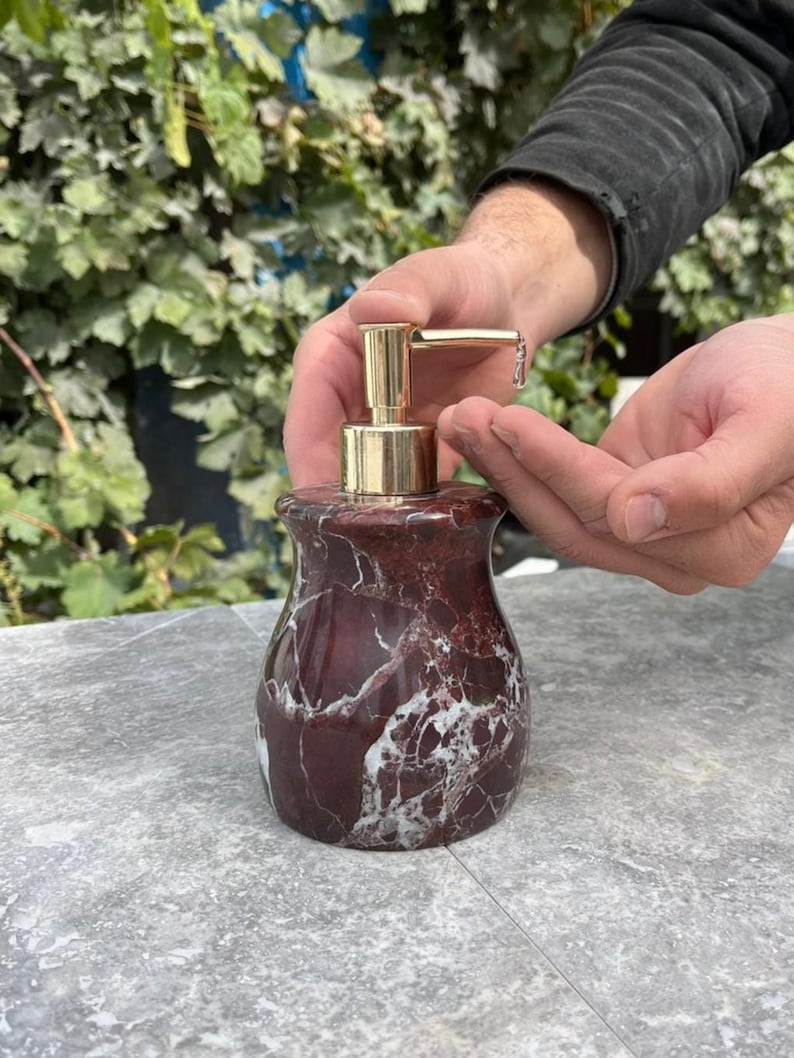 Puede incluir: Un dispensador de jab&oacute;n de m&aacute;rmol con una bomba dorada. El dispensador es de color rojo oscuro con vetas blancas. El dispensador est&aacute; sobre una superficie gris.