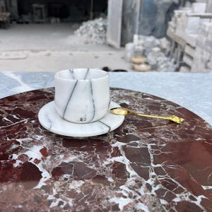 Op de afbeelding: Een witte marmeren kop en schotel met zwarte adering, op een rood en wit marmeren oppervlak. Een kleine gouden lepel ligt op de schotel. De set is ontworpen voor het serveren van thee of koffie.