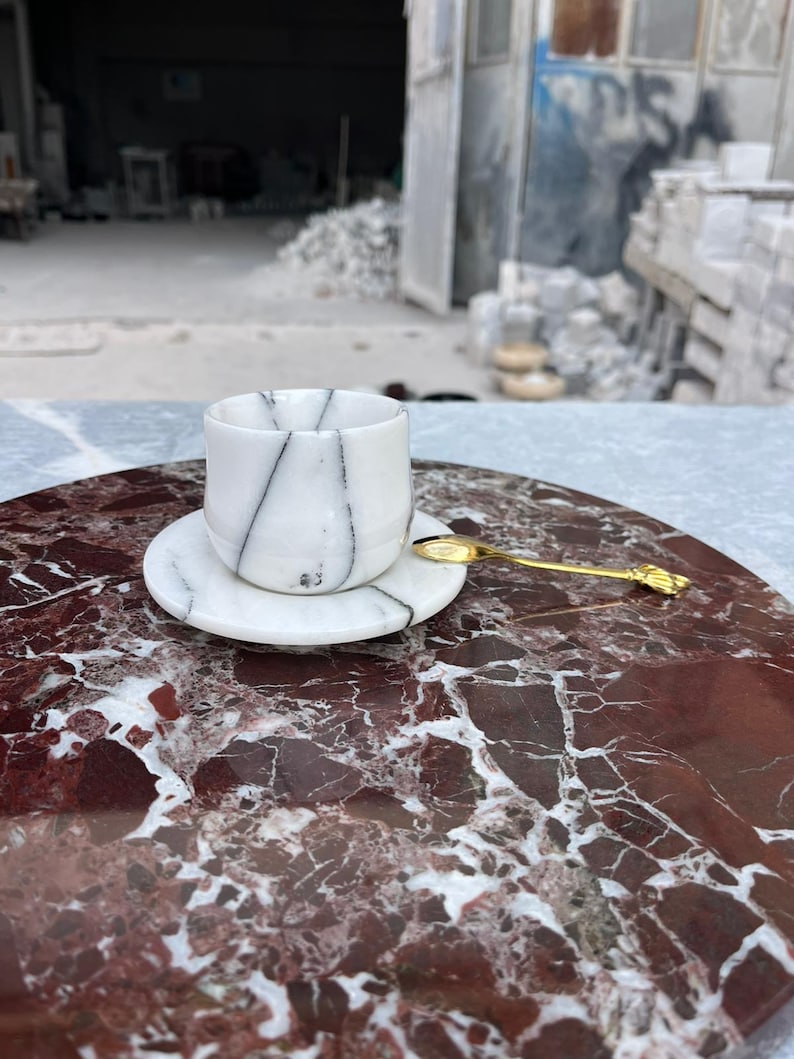 Op de afbeelding: Een witte marmeren kop en schotel met zwarte adering, op een rood en wit marmeren oppervlak. Een kleine gouden lepel ligt op de schotel. De set is ontworpen voor het serveren van thee of koffie.