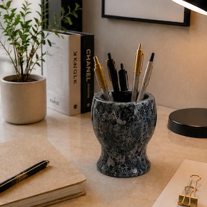 May include: A cylindrical pen holder with a dark grey and white marbled pattern, containing several pens with gold and silver accents. The holder has a flared base and a wide opening, sitting on a light-coloured surface alongside a notebook and pens.