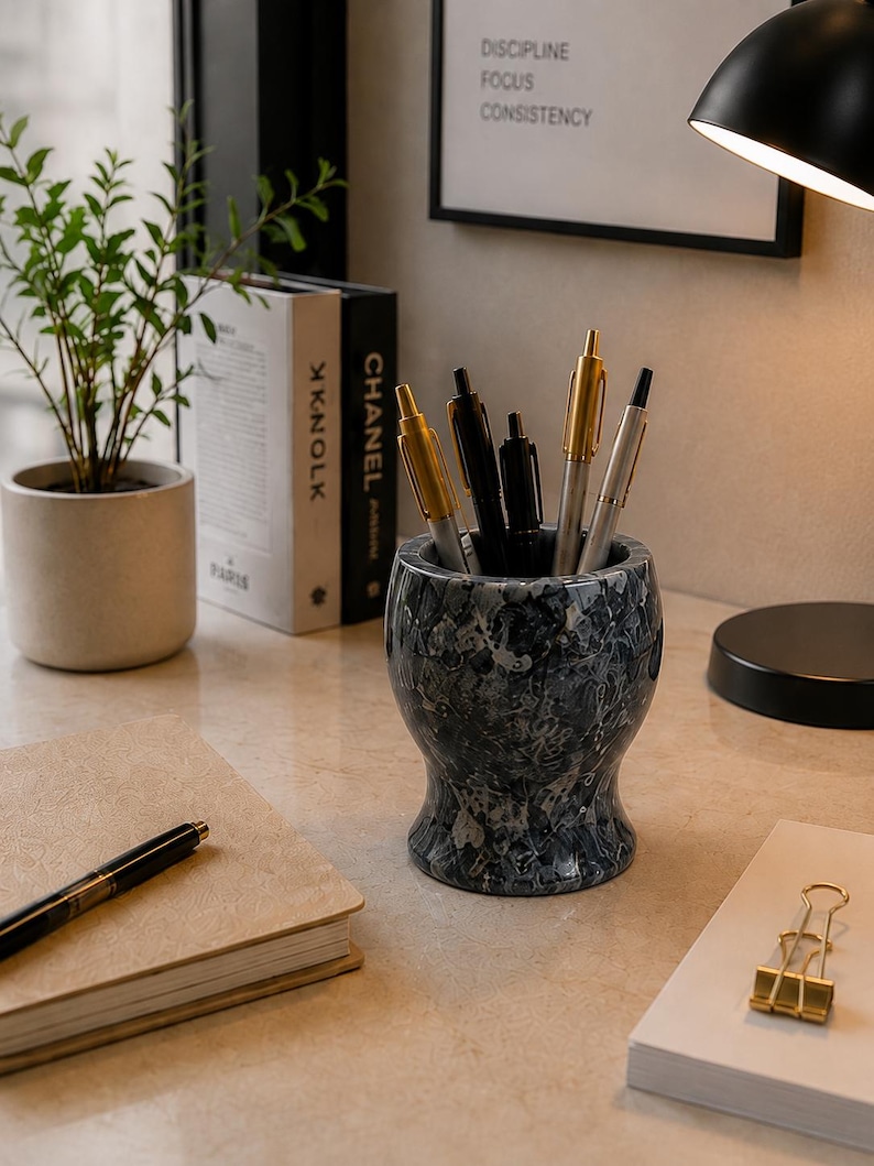 May include: A cylindrical pen holder with a dark grey and white marbled pattern, containing several pens with gold and silver accents. The holder has a flared base and a wide opening, sitting on a light-coloured surface alongside a notebook and pens.
