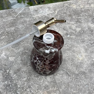 Puede incluir: Un dispensador de jab&oacute;n decorativo con un patr&oacute;n similar al m&aacute;rmol en tonos burdeos y blanco. El dispensador tiene una bomba dorada y un tubo de pl&aacute;stico transparente. La bomba est&aacute; desenroscada, revelando una abertura de pl&aacute;stico blanco.